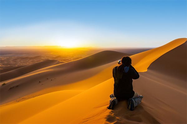 Handsome man with dreadlocks taking a photograph of sunset from a sand dune By Andrea Obzerova